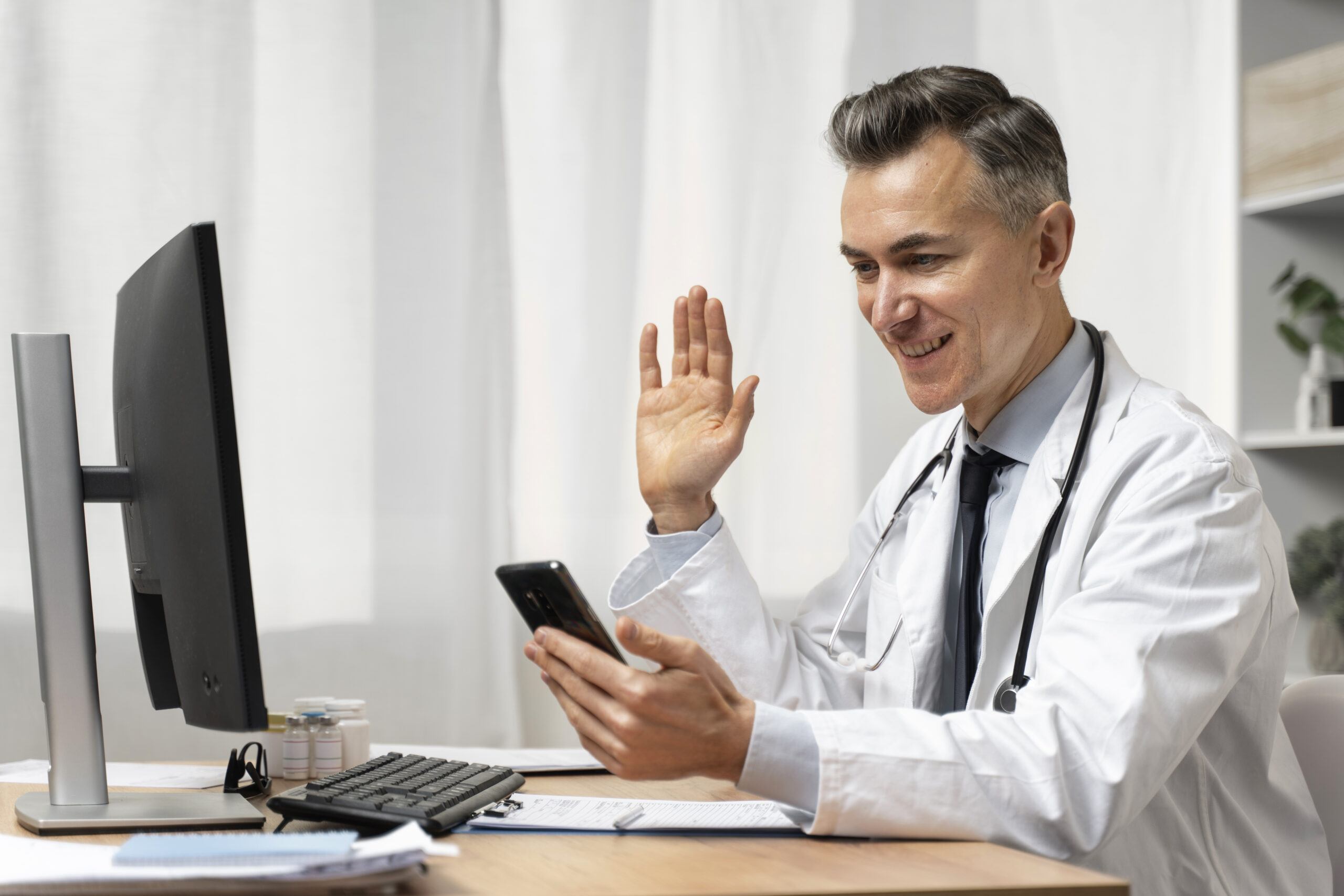 A doctor is waving at his phone while sitting in front of a computer.